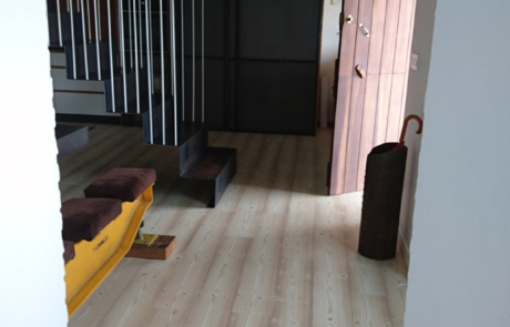 pasillo de una vivienda, se ve la puerta de salida entreabierta y en frente de ella las escaleras de acceso al piso superior