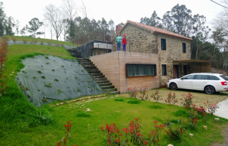 dos niños desde la terraza ajardinada superior de una vivienda, se ve la fachada de piedra y dos coches aparcados en la entrada